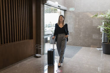 Smiling businesswoman with suitcase walking into hotel - JRVF03038
