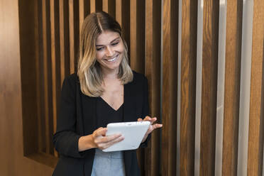 Smiling businesswoman using tablet PC standing by wooden wall at hotel - JRVF03046