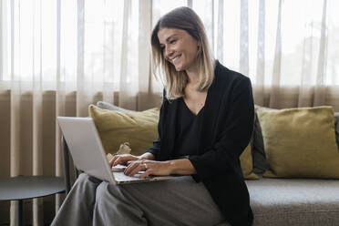 Smiling businesswoman using laptop sitting on sofa in lobby - JRVF03058
