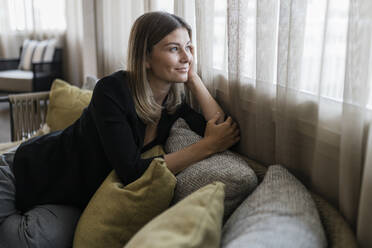 Lächelnde Geschäftsfrau, die durch ein Fenster schaut und auf einem Sofa im Hotel sitzt - JRVF03062