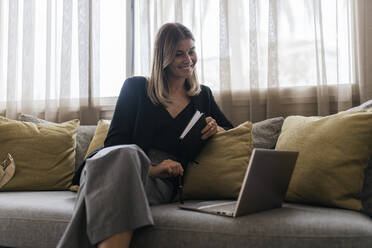 Lächelnde Geschäftsfrau, die eine Molkerei hält und auf einen Laptop schaut, der auf einem Sofa steht - JRVF03065
