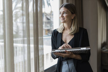 Glückliche Geschäftsfrau mit Laptop, die durch das Fenster eines Hotels schaut - JRVF03071