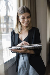 Smiling businesswoman using laptop standing by window at hotel - JRVF03072