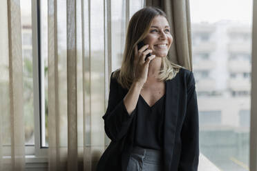 Smiling businesswoman talking on phone standing by window - JRVF03078