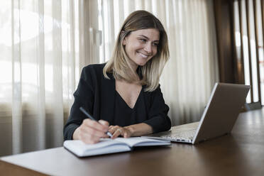 Lächelnde Geschäftsfrau, die auf einen Laptop-Bildschirm schaut und in ein Tagebuch auf einem Tisch schreibt - JRVF03085