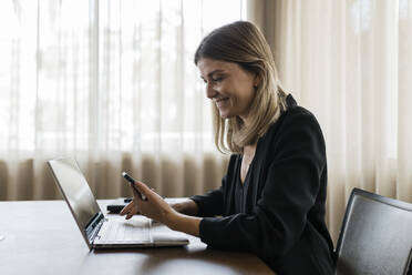 Smiling businesswoman holding mobile phone sitting with laptop on table - JRVF03087