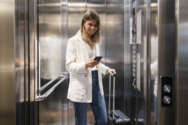 Smiling businesswoman using smart phone standing with suitcase in elevator - JRVF03091