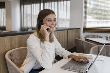 Smiling businesswoman talking on mobile phone sitting with laptop at restaurant - JRVF03096