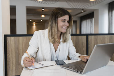 Lächelnde Geschäftsfrau, die im Restaurant auf einen Laptop-Bildschirm schaut und in ihr Tagebuch schreibt - JRVF03097