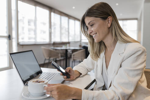 Lächelnde Geschäftsfrau mit Mobiltelefon und Kaffeetasse in einem Café sitzend - JRVF03101