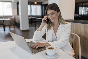 Smiling businesswoman talking on phone sitting with laptop at cafe - JRVF03102