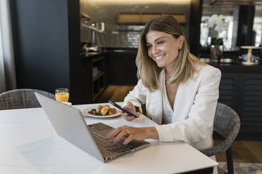 Smiling businesswoman holding mobile phone using laptop at restaurant - JRVF03103