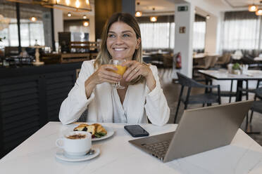 Lächelnde Geschäftsfrau mit einem Saftglas in der Hand und einem Laptop im Restaurant - JRVF03104