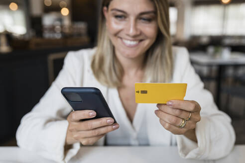 Smiling businesswoman holding credit card doing online shopping through smart phone at restaurant - JRVF03117