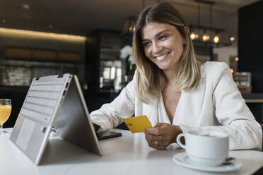 Happy businesswoman holding credit card doing online shopping through laptop at restaurant - JRVF03119