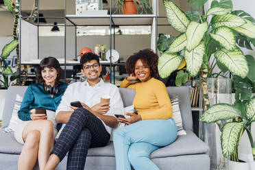 Lächelnde multirassische Kollegen mit Smartphones auf dem Sofa im Büro - MEUF06720