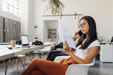 Businesswoman with documents talking on smart phone in office - MEUF06732