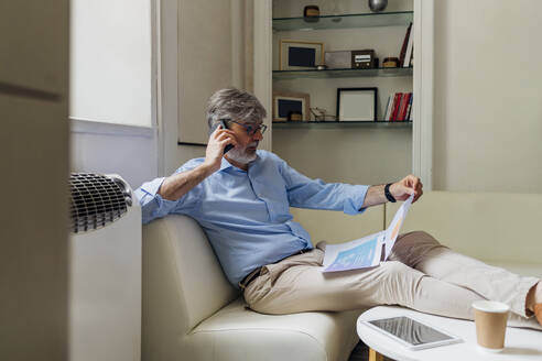 Älterer Geschäftsmann mit Dokumenten im Gespräch auf dem Smartphone im Büro - MEUF06741