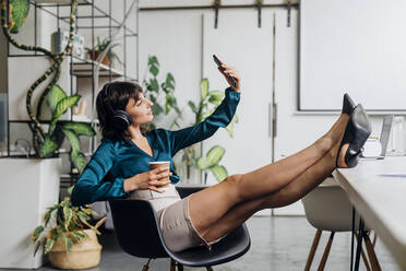 Businesswoman with headphones taking selfie on smart phone at workplace - MEUF06751