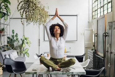 Young businesswoman doing yoga with hands clasped on desk in office - MEUF06753