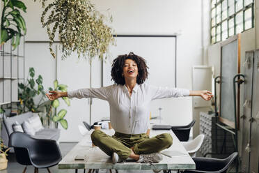 Happy businesswoman sitting with arms outstretched on desk at office - MEUF06754