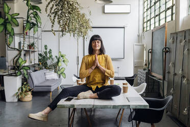 Businesswoman meditating with hands clasped on desk at workplace - MEUF06762