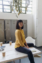 Smiling businesswoman sitting on desk at workplace - MEUF06764