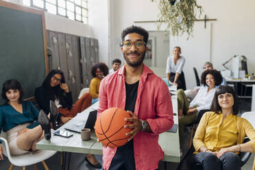 Smiling businessman with basketball by multiracial colleagues in office - MEUF06772