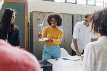 Smiling businesswoman showing wind turbine model to colleagues in office - MEUF06778