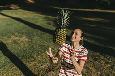 Glückliche Frau spielt mit Ananas im Park an einem sonnigen Tag - DMGF00759