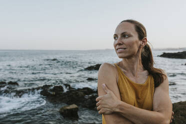 Glückliche Frau umarmt sich selbst am Meeresufer bei Sonnenuntergang - DMGF00771