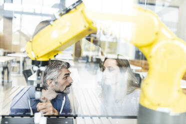 Engineers pointing at industrial robot in glass talking to colleague - WESTF24847