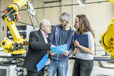 Manager discussing technical report with coworkers in front of industrial robot - WESTF24871