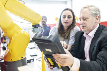 Senior engineer looking at digital control of female coworker at industrial robot - WESTF24954