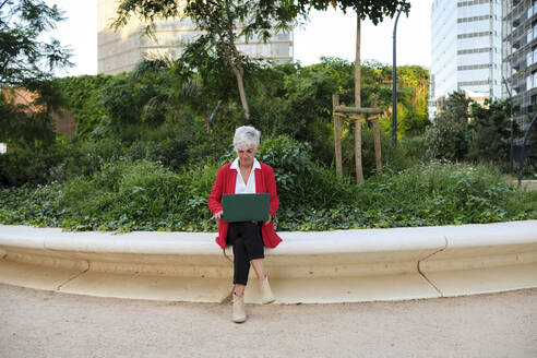 Mature businesswoman using laptop sitting on wall - AGOF00301