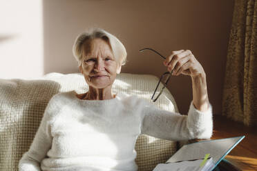 Smiling senior woman holding eyeglasses sitting on sofa at home - OSF00202