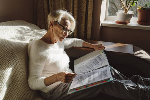 Woman wearing eyeglasses reading documents sitting in living room at home - OSF00206