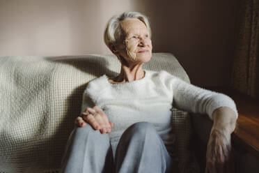 Smiling senior woman with white hair sitting on sofa at home - OSF00210