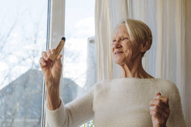 Glückliche ältere Frau mit Palo-Santo-Stock am Fenster - OSF00223