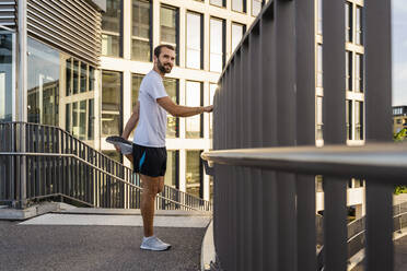 Smiling man stretching leg standing by railing on walkway - DIGF18203