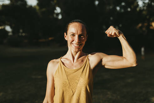 Happy woman flexing biceps in park - DMGF00778