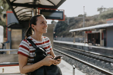 Smiling woman with smart phone standing at railroad station - DMGF00792