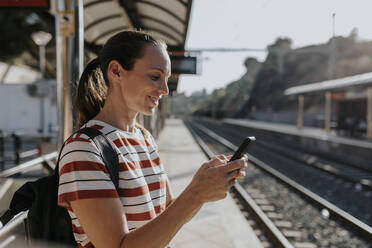 Smiling woman using smart phone standing at railroad station - DMGF00795