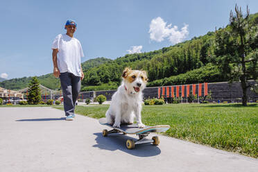 Smiling man with dog sitting on skateboard in park - OMIF00954