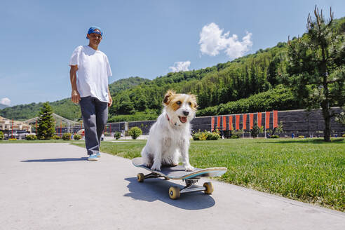 Lächelnder Mann mit Hund auf einem Skateboard im Park sitzend - OMIF00954