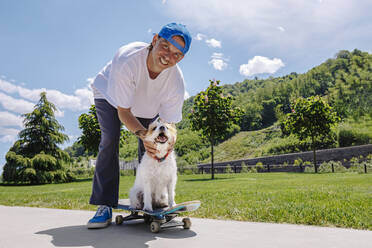 Glücklicher Mann mit Hund auf Skateboard im Park sitzend - OMIF00958