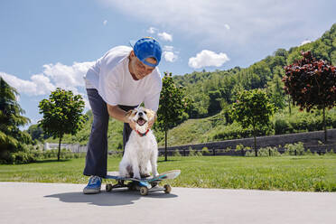 Lächelnder Mann streichelt Hund, der auf einem Skateboard sitzt - OMIF00959