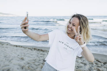 Happy woman gesturing peace sign talking selfie through smart phone at beach - SIF00241