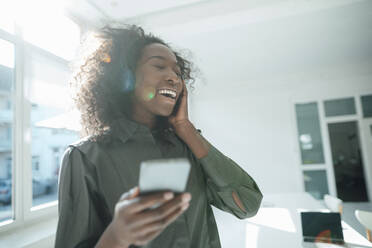 Happy young woman holding mobile phone listening music through wireless headphones - KNSF09470