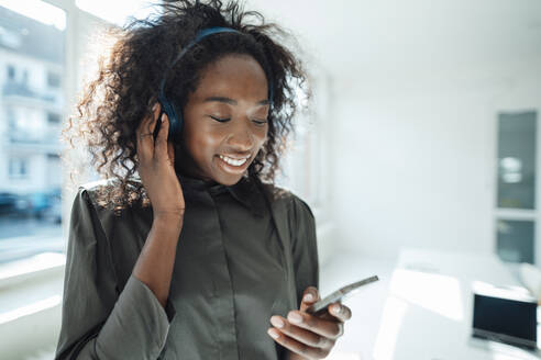 Lächelnde junge Frau, die ein Smartphone benutzt und über drahtlose Kopfhörer Musik hört - KNSF09472